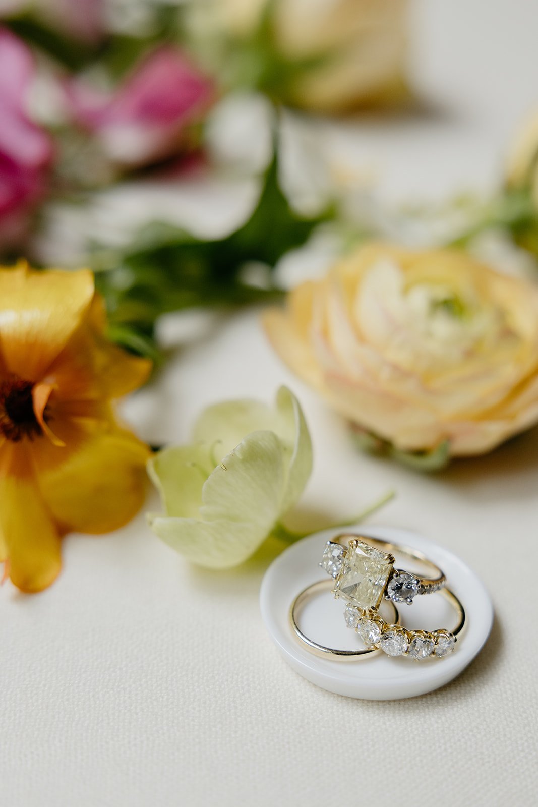 3 gold wedding rings, 1 wedding band and two diamond bands with a yellow center diamond on a small tray with flowers surrounding it.