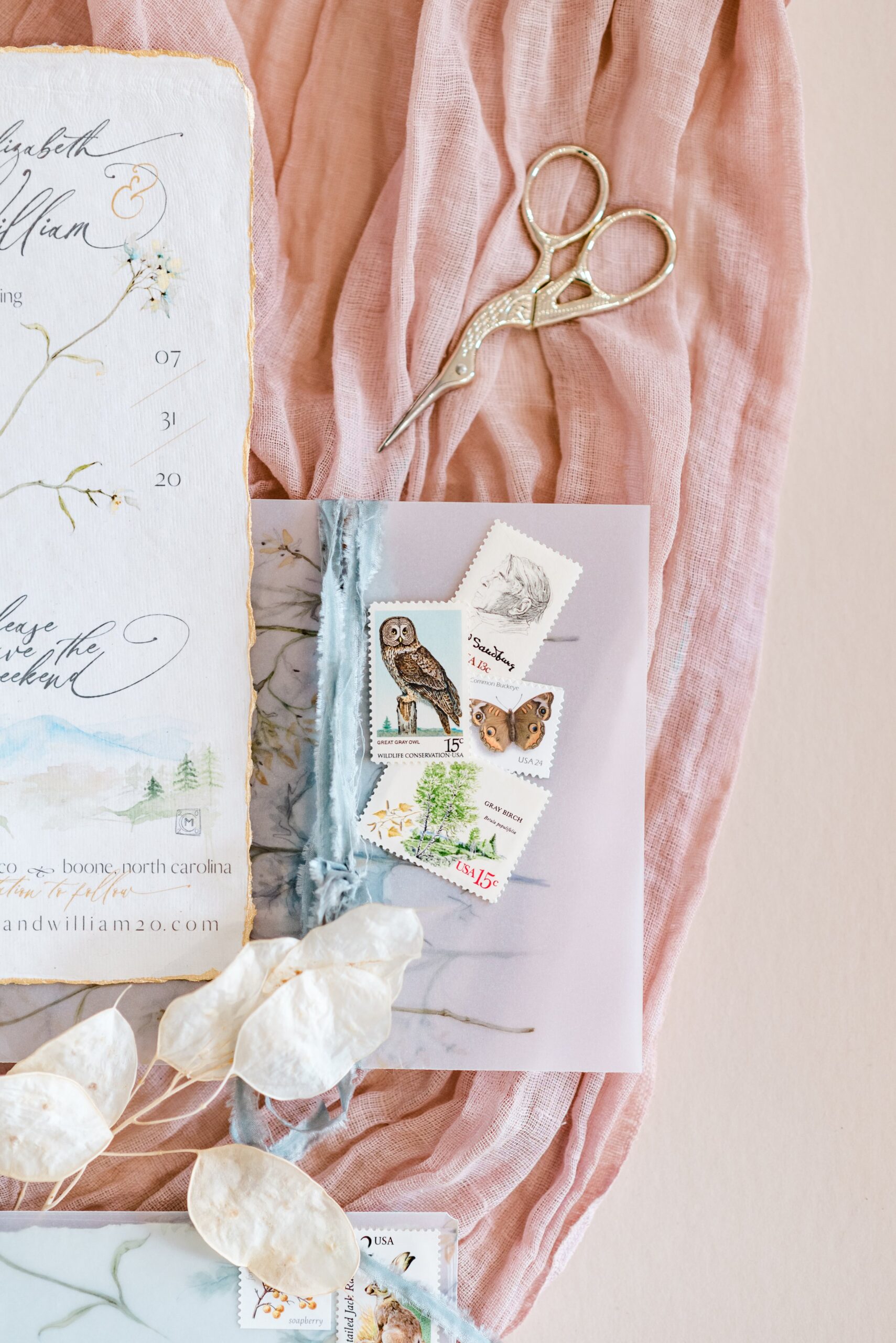 A flatlay on a pink linen with details including stationery, mini gold scissors and vintage stamps featuring wildlife. 