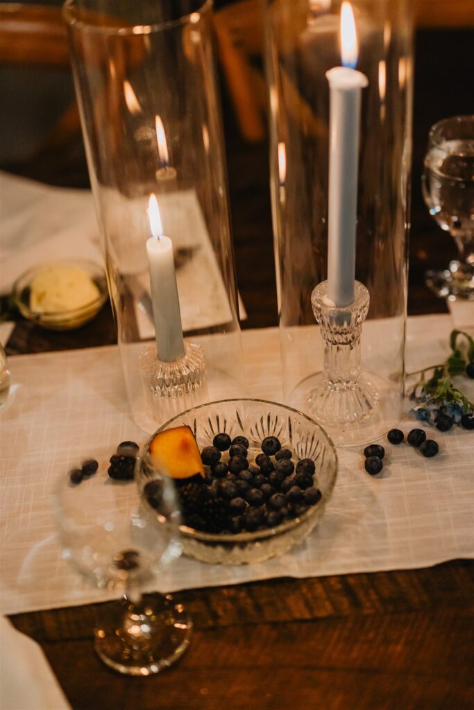 Wedding tablescape on a brown farmstyle table with candles, glassware, and blueberries in a bowl.